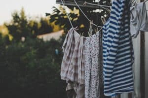 Clothing hangs on a washing line outdoors, including a striped shirt and floral-patterned garments, set against green trees and a sunny sky in the blurred background.