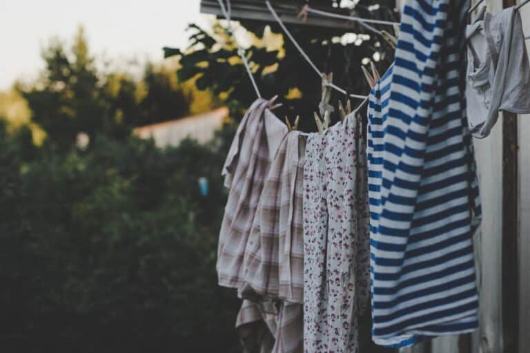 Clothing hangs on a washing line outdoors, including a striped shirt and floral-patterned garments, set against green trees and a sunny sky in the blurred background.