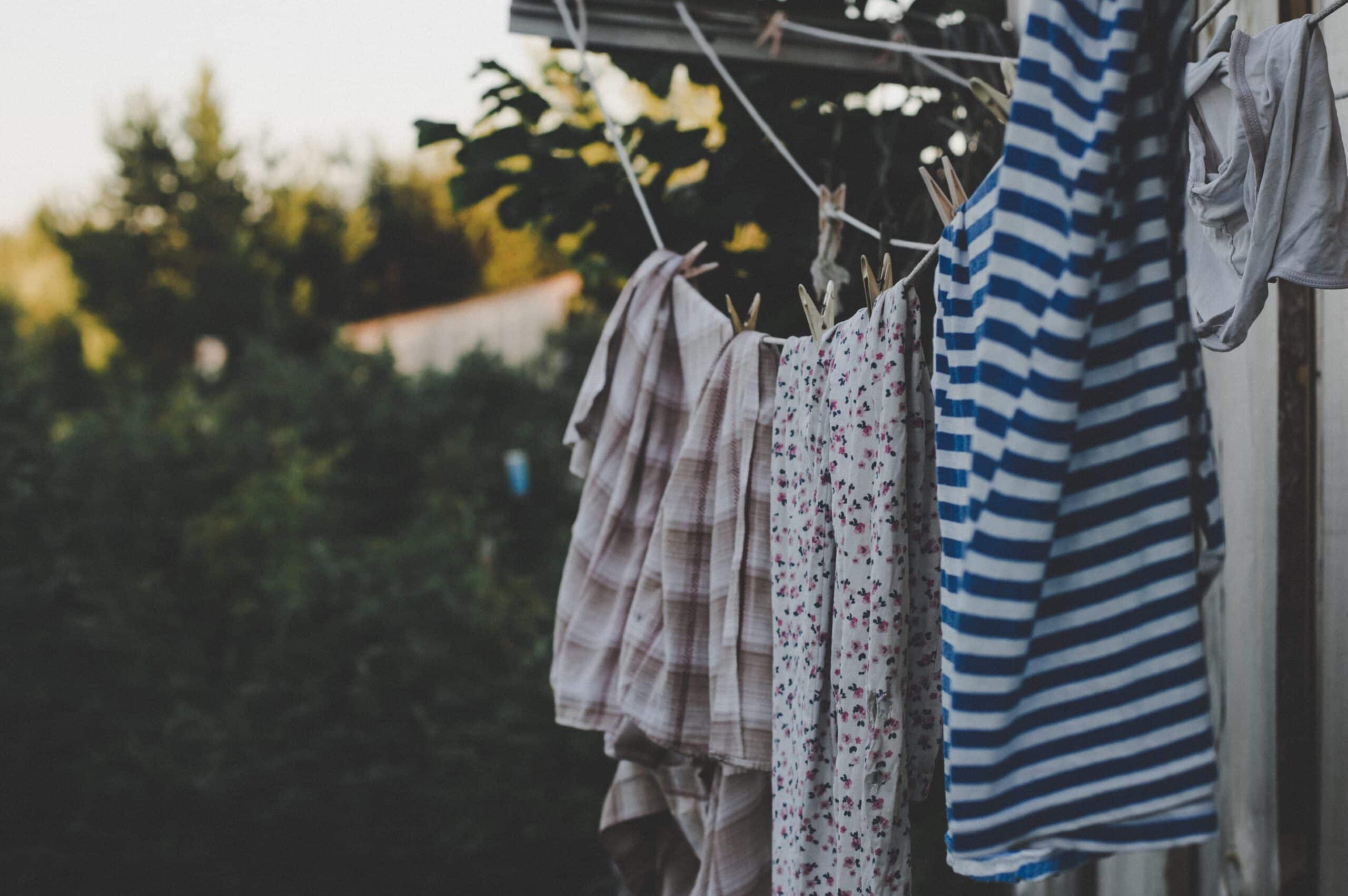 Clothing hangs on a washing line outdoors, including a striped shirt and floral-patterned garments, set against green trees and a sunny sky in the blurred background.