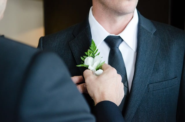 A person in a suit adjusts a buttonhole made of white flowers and green leaves on the lapel of another man, highlighting his best man's outfit and classic wedding suit.