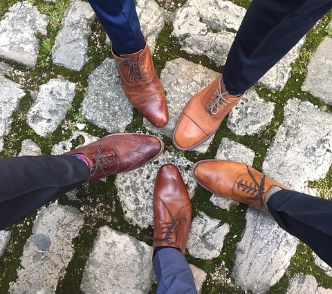 Four people wearing different shades of brown dress shoes and dark trousers—ideal inspiration for best man attire—stand in a circle on a cobblestone path with moss growing between the stones.