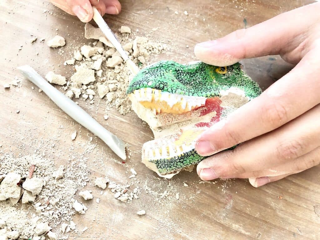 Close-up of hands using a small tool to excavate a toy dinosaur skull from a block of plaster—dust and debris scattered—as part of a National Geographic STEM Dig Kit.