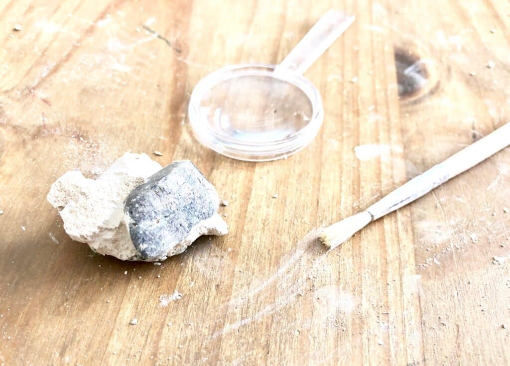 A magnifying glass, a small brush, and a partially unearthed rock or fossil from a National Geographic STEM Dig Kit are placed on a wooden surface covered in dust and debris.
