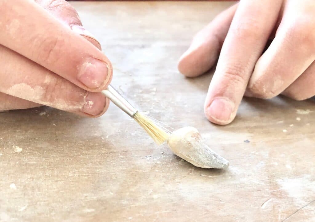 A close-up of hands brushing dust off a small fossil or artefact on a wooden surface using a fine paintbrush, similar to the experience offered by National Geographic STEM Dig Kits.