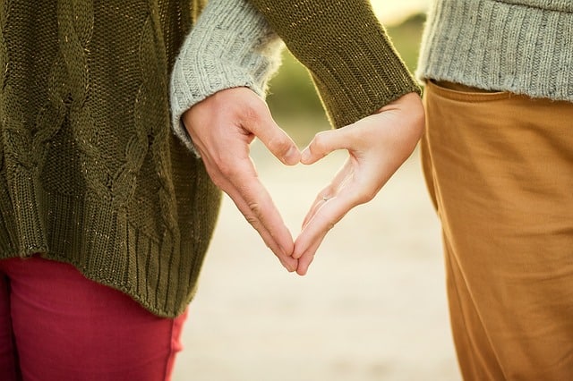 Two people stand side by side, each wearing jumpers. They form a heart shape with their hands together, symbolising love and connection—capturing a moment of relationship repair and hope for those looking to mend bonds. Only their torsos, arms, and hands are visible.