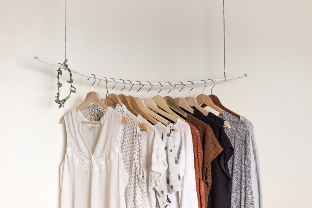 A minimalist clothes rail made from a white branch hangs from the ceiling, displaying femmeluxe neutral-toned tops and dresses on wooden hangers against a plain white wall.
