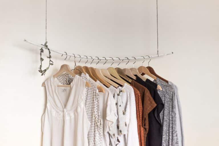 A minimalist clothes rail made from a white branch hangs from the ceiling, displaying femmeluxe neutral-toned tops and dresses on wooden hangers against a plain white wall.