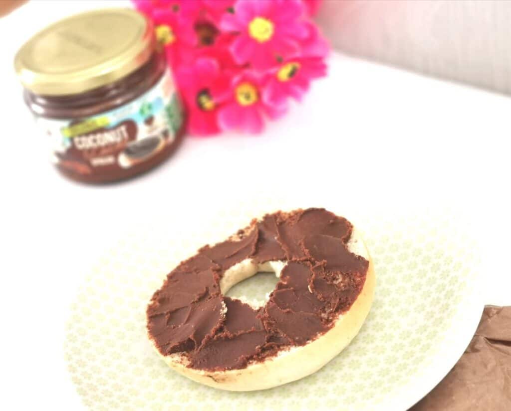 A sliced bagel topped with chocolate spread sits on a patterned plate. In the background, thereβs a jar of chocolate spread, a bunch of bright pink flowers, and hints of coconut for new ways to use coconut in your breakfast routine.