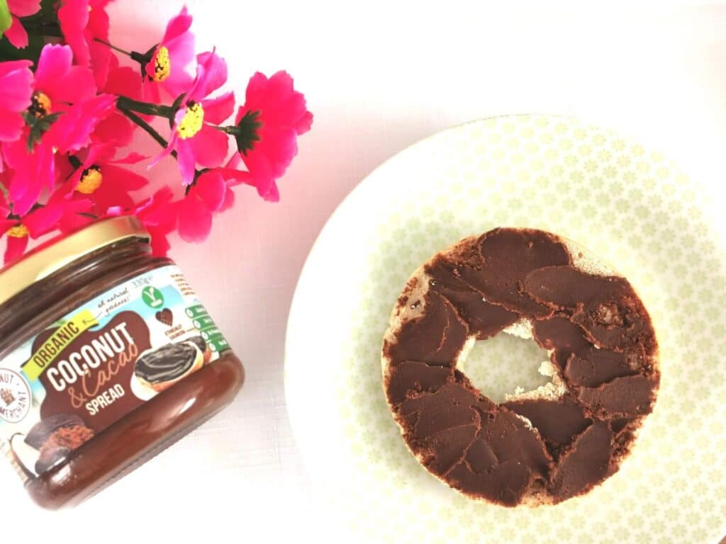 An open jar of organic coconut cocoa spread sits next to a bagel half topped with the delicious spread. Pink artificial flowers add a touch of charm, hinting at the different ways you can enjoy this treat or even win it in a competition.