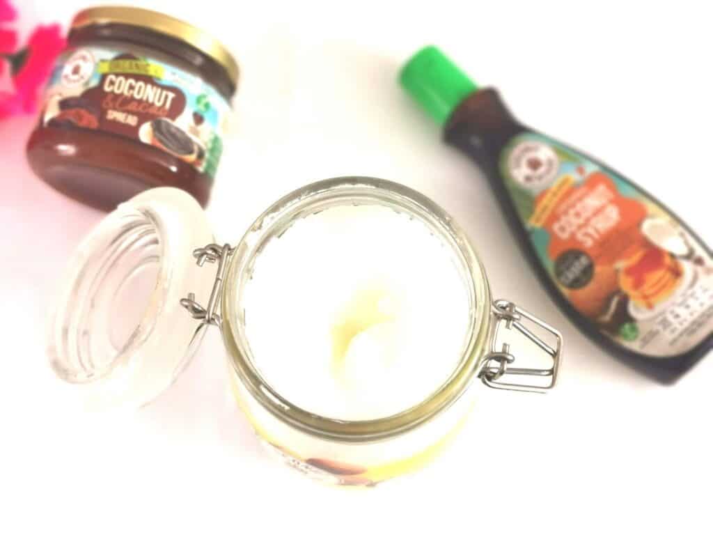 An open glass jar filled with white coconut oil is in the centre, with a jar of coconut spread and a bottle of coconut syrup in the backgroundβshowcasing versatile coconut uses on a clean white surface.