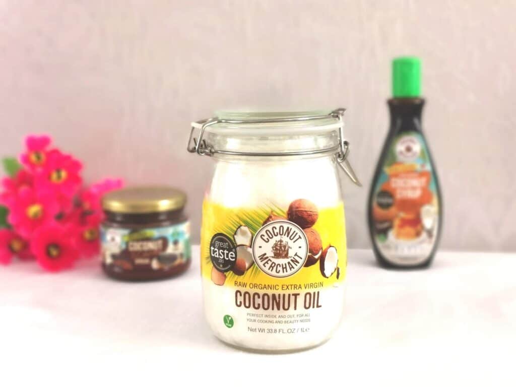A jar of organic extra virgin coconut oil is centred on a white surface. In the background, a jar of chocolate spread, a bottle of syrup, and pink flowers are softly blurredβperfect inspiration for creative coconut uses or your next competition.