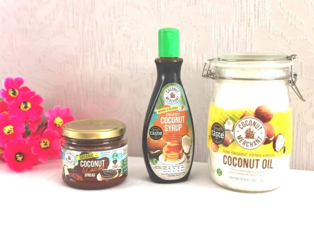 Three coconut productsβcoconut oil in a glass jar, coconut syrup in a squeezy bottle, and coconut chocolate spreadβare displayed on a white surface with pink artificial flowers, highlighting creative coconut uses perfect for any Coconut Giveaway.