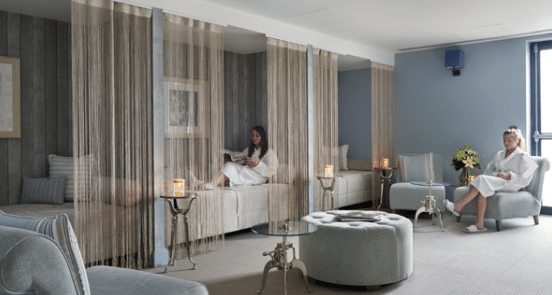 A modern spa lounge with soft blue-grey decor, string curtains dividing seating areas—a perfect spa day for two. Two women in white robes relax, one reading on a daybed, the other beside flowers and lit candles on small tables.