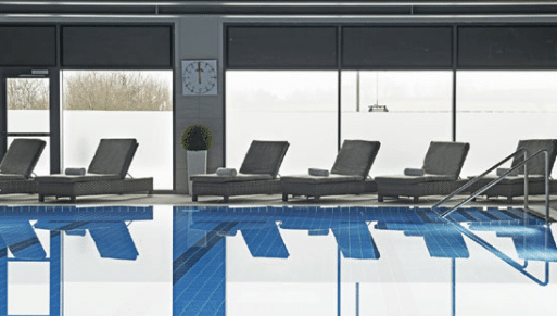 Indoor swimming pool with clear blue water, several empty loungers, large windows letting in natural light, a potted plant, and a clock on the wall—a perfect spa day win for two with snowy scenery visible outside.
