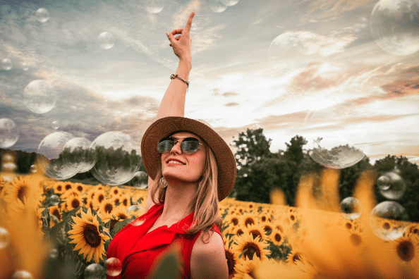 A smiling woman wearing sunglasses and a hat stands in a sunflower field, pointing up at bubbles floating in the air under a dramatic sky—embracing personal growth and coping with change amidst nature’s beauty.