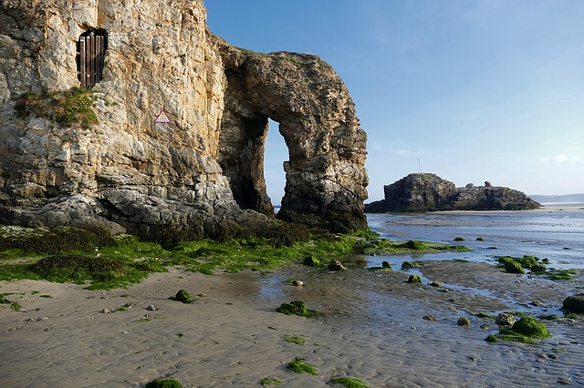 A natural rock arch stands on a sandy, seaweed-covered beach under a blue sky—an intriguing spot for a fun break with children. A barred window and warning sign are set in the rock, with tidal water and an island in the background.
