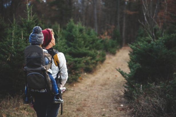 A person enjoying a family holiday walks along a dirt path in a forested area, carrying a child in a rucksack carrier. Both are bundled up in hats and jackets, surrounded by evergreen trees.