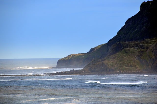 Waves roll towards a rocky East Coast shoreline with steep, green cliffs under a clear blue sky. The sea appears slightly misty near the shore, and the landscape looks perfect for a peaceful weekend getaway.