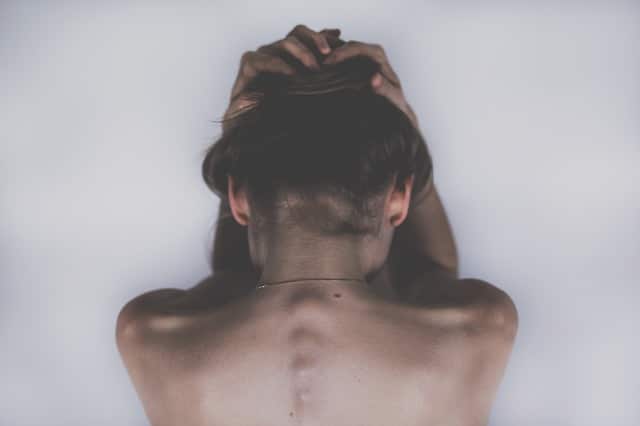 A person with short hair is seen from behind, sitting with their head bowed and hands clasped over the top of their head, exposing their bare back and spine—a moment capturing the tension of recovering from back injury.