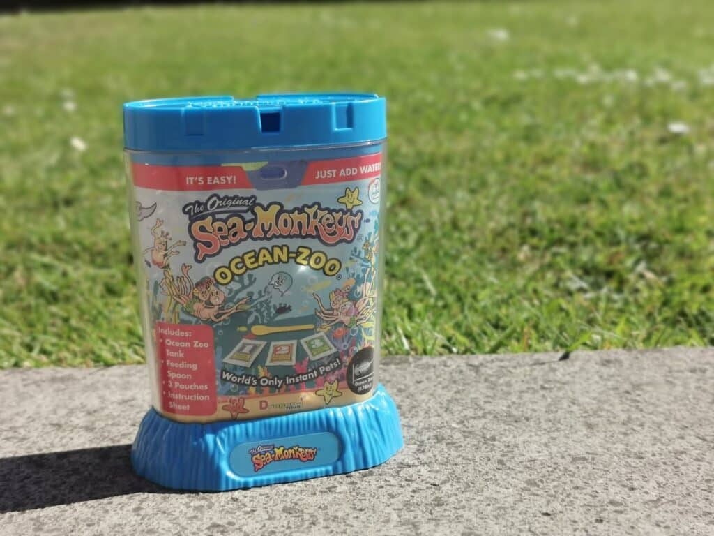 A Sea Monkeys Ocean-Zoo kit sits on a concrete surface with green grass in the background. The blue container features colourful, cartoonish aquarium graphics and instructions—perfect for fans of Bandai STEM Kits and hands-on learning fun.