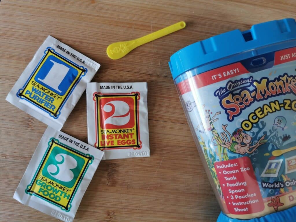 Packets labelled "1 Water Purifier," "2 Sea-Monkey Eggs," and "3 Growth Food" sit on a wooden surface beside a yellow spoon and a colourful Sea Monkeys Ocean-Zoo tank, perfect for fans of hands-on fun like Crystal Growing or Bandai STEM Kits.