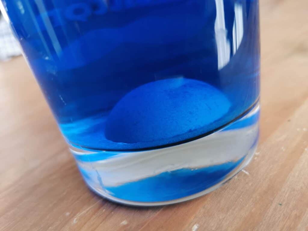 A blue-dyed egg sits at the bottom of a clear glass filled with blue liquid, resembling the mesmerising hues seen in Crystal Growing kits, and rests on a wooden surface.