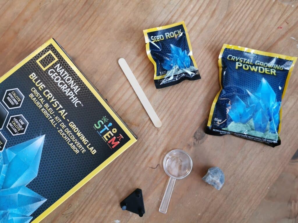 A National Geographic Blue Crystal Growing Lab kit, part of the Bandai STEM Kits range, is spread out on a wooden surface with a booklet, "Seed Rock," "Crystal Growing Powder," wooden stick, plastic magnifier, and a rock.