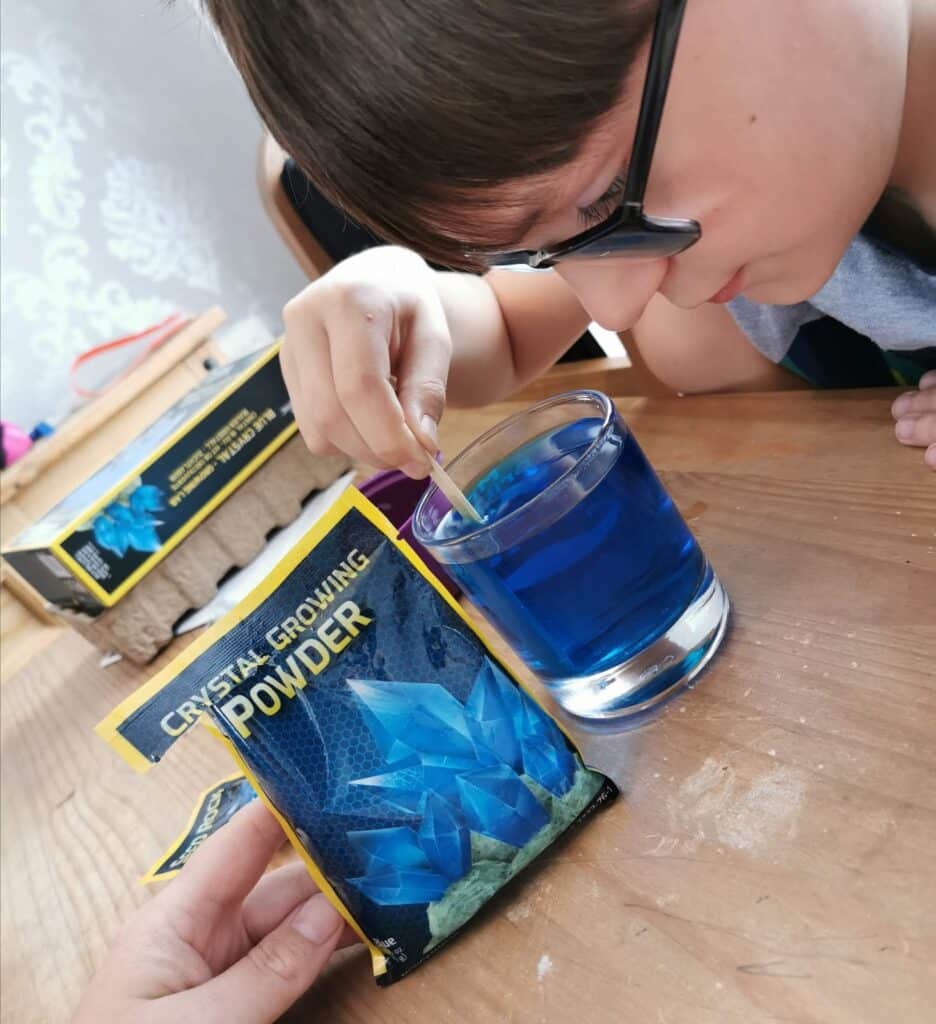 A child wearing glasses uses a stick to stir a glass of blue liquid on a wooden table, next to a packet labelled "Crystal Growing Powder." Crystal Growing kits and other materials are visible in the background.