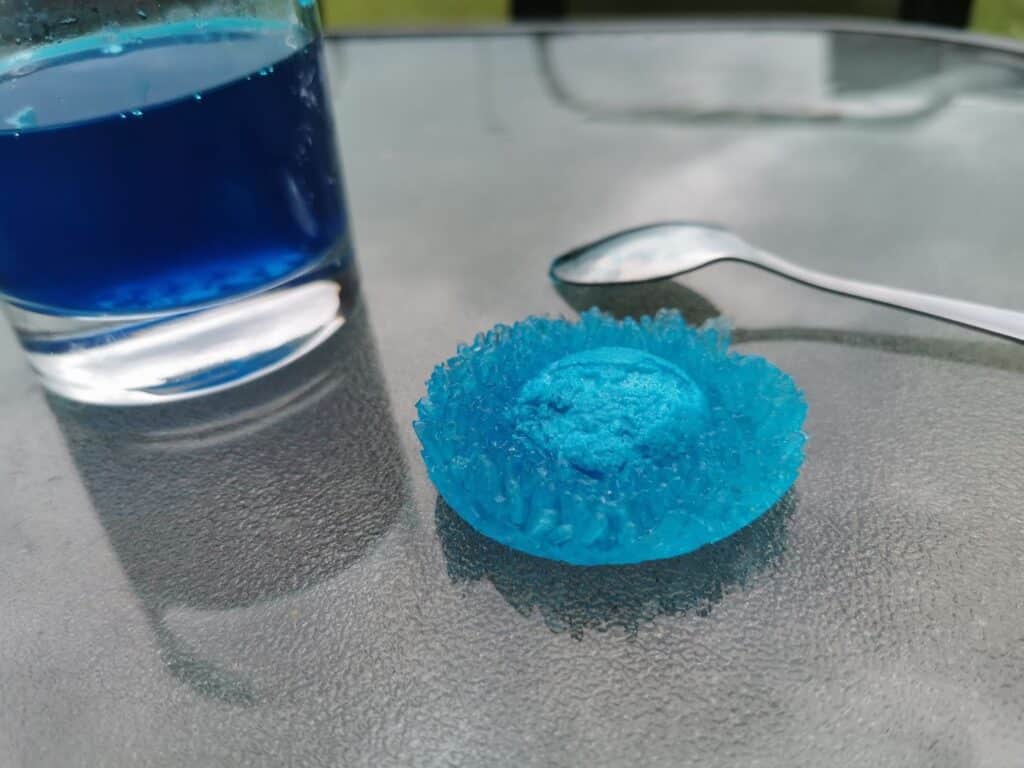 A round, crystalline blue sweet sits on a glass table next to a spoon and a glass filled with blue liquid, its surface sparkling like Crystal Growing kits and giving the scene a cool, refreshing feel.
