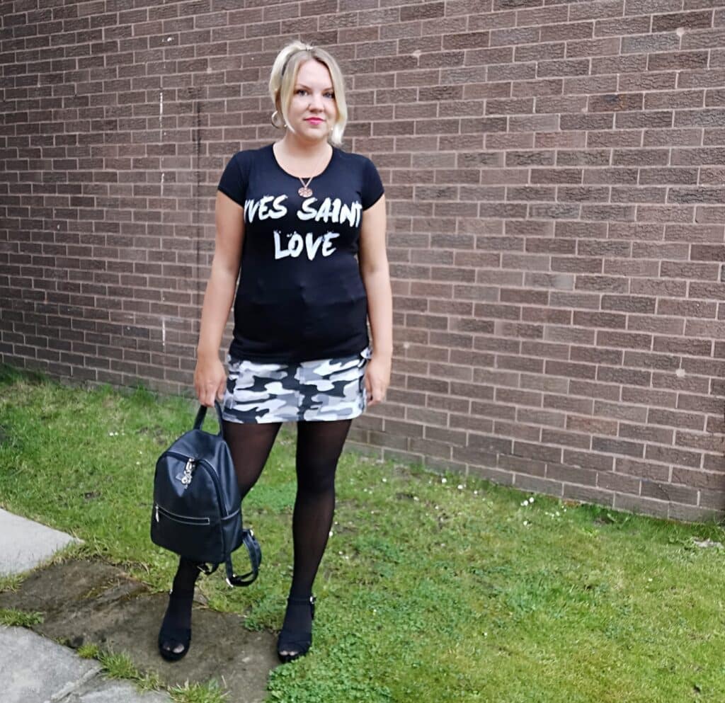 A woman stands outdoors on grass by a brick wall, wearing a black "YES SAIN LOVE" T-shirt and holding a black rucksack—ready to win an Amazon Voucher worth £20 with her chic look. She sports a camouflage skirt, black tights, heeled shoes, and blonde hair.