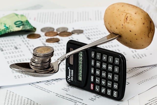 A potato and a spoon with stacked coins are balanced on a calculator, surrounded by scattered papers, coins, and a banknote, symbolising family savings and financial balance.