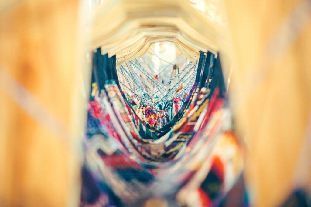 A close-up, blurred view of colourful patterned clothes hanging on wooden hangers, creating a tunnel-like visual effect with vibrant femmeluxe fabrics in focus at the centre.