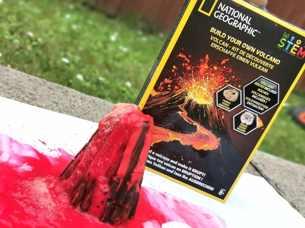 A homemade volcano model erupts with red liquid on white paper, next to a National Geographic "Build Your Own Volcano" science kit box from the Bandai STEM Kits collection, outdoors on grass.