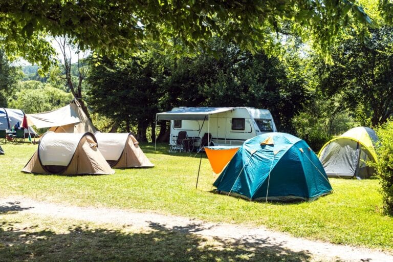 A sunny camping campsite with several colourful tents and a white camper van set up on green grass, surrounded by trees. Folding chairs and tables are arranged near the camper under the shade.