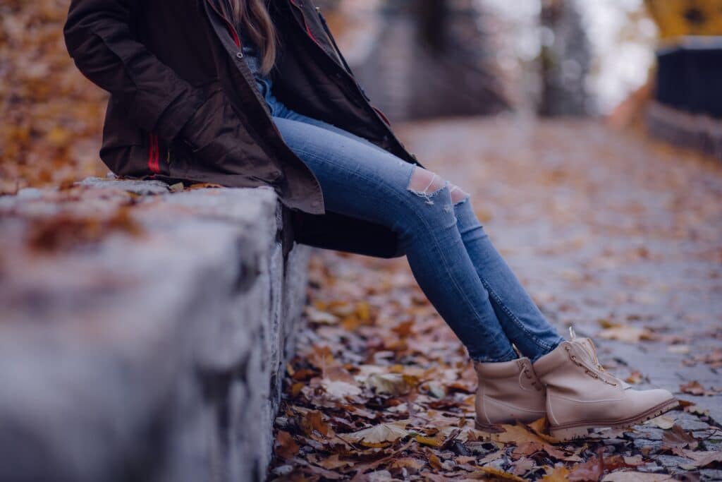 A person dressing down in a dark jacket, ripped blue jeans, and light tan boots sits on a stone wall surrounded by autumn leaves on a path. Their face is not visible.