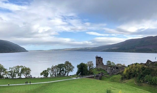 A scenic view of a calm lake with rolling hills in the background, lush green grass in the foreground, and the ruins of an old stone castle by the water’s edge—a perfect spot for family walks and child-friendly strolls under a partly cloudy sky.