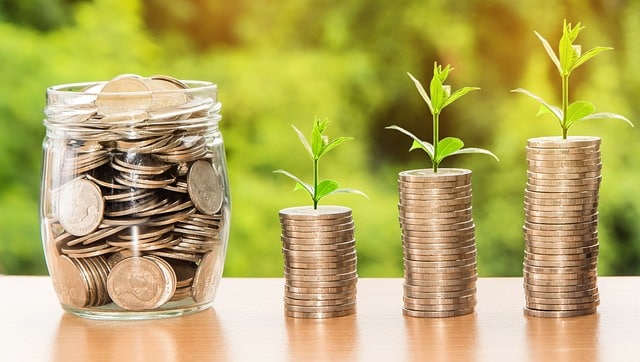 A glass jar filled with coins sits next to three stacks of coins, each topped with a small green plant sprout, symbolising saving for your dream home and smart investment against a blurred green background.