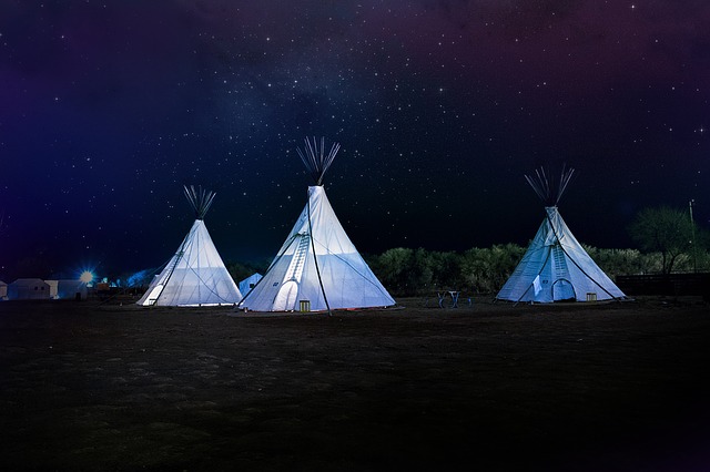 Three illuminated tipis stand in a row under a clear, starry night sky, creating a magical camping scene surrounded by trees and shadows on the dark, open ground.
