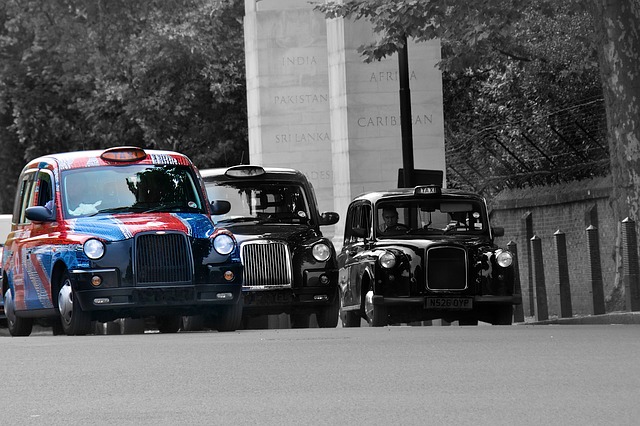 Three London taxis—two black and one decorated with a colourful pattern—drive on a city street near a large stone monument. Trees line the road and metal posts mark the pavement, offering travellers a look at cheap taxis while exploring the city’s sights.