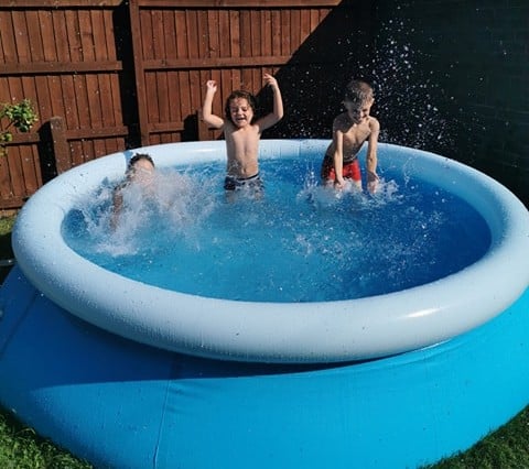 Three children play and splash in a blue inflatable paddling pool outdoors on a sunny day—perfect for Outdoor Party Ideas. One child stands with arms raised, while others create waves and droplets. A wooden fence sits in the background.
