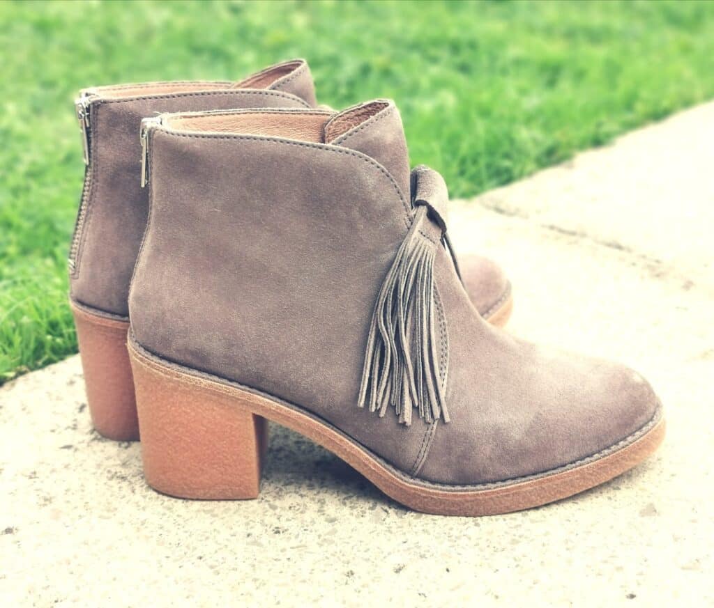 A pair of taupe suede ankle boots with chunky heels, zip backs, and decorative fringe tassels—these stylish shoes offer a Unique UGG look, breaking away from the Not Classic style you might expect, placed on a pavement near green grass.