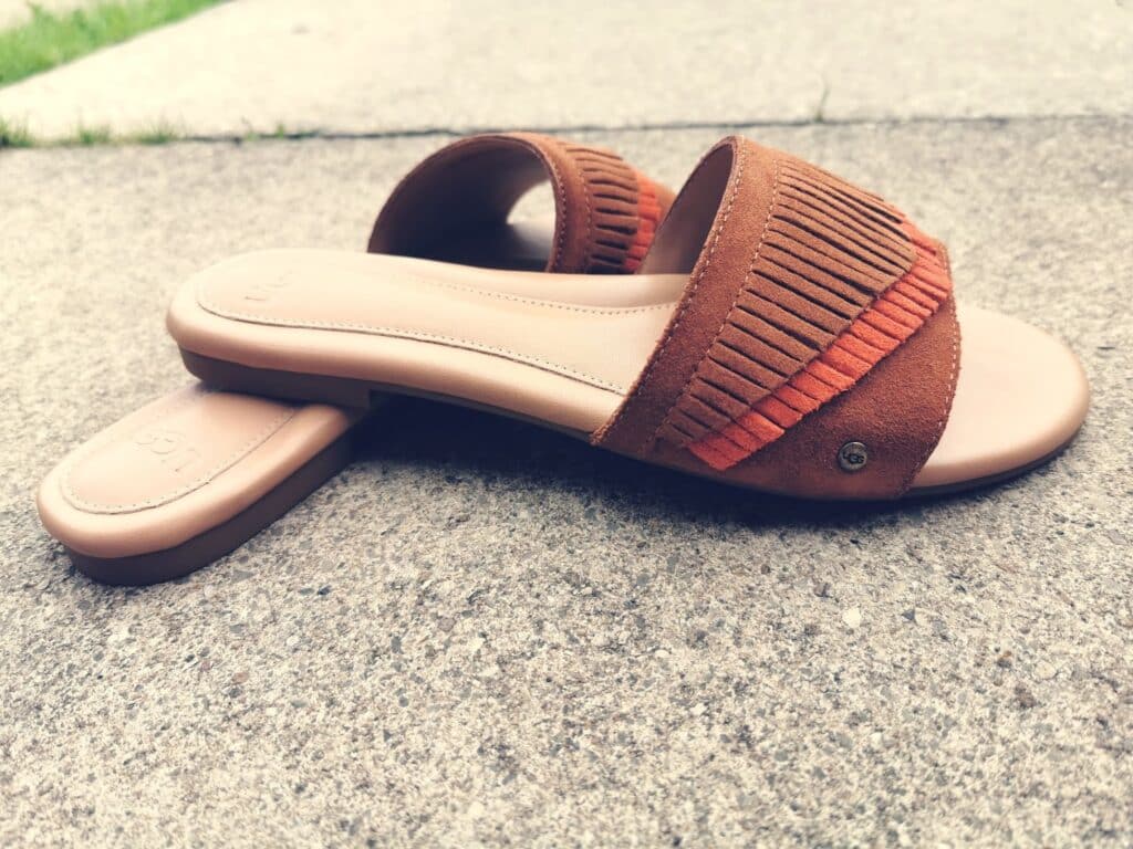 A pair of brown suede slide sandals with fringe detail on the straps, placed on a concrete surface. Not classic like UGG boots, one sandal lies flat whilst the other is propped on its side.