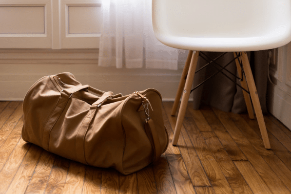 A brown holdall sits on a wooden floor next to a white chair with wooden legs—ready for summer travel in a room with white panelled walls and sheer curtains.
