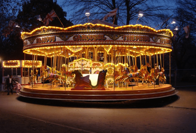 A brightly lit, vintage carousel with colourful horses spins at dusk in a park. The warm lights contrast with the darkening sky and silhouetted trees, making it a perfect spot for outdoor party ideas or a magical children's party.