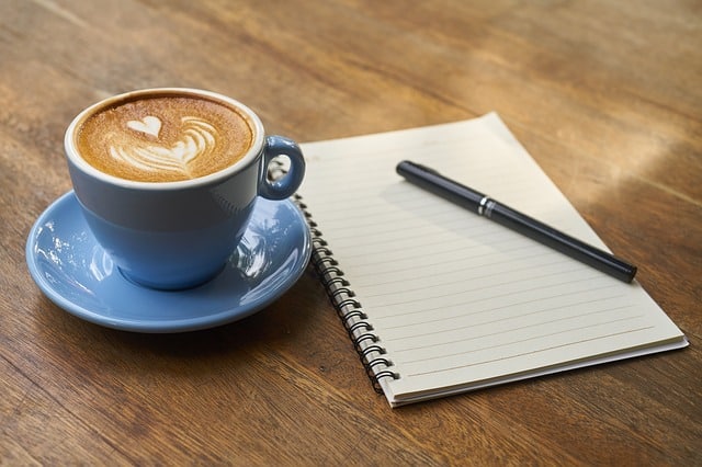A blue cup of coffee with heart-shaped latte art sits on a saucer next to an open, lined spiral notebook, ready for jotting down 4 ways to successfully save more money, along with a black pen on a wooden table.