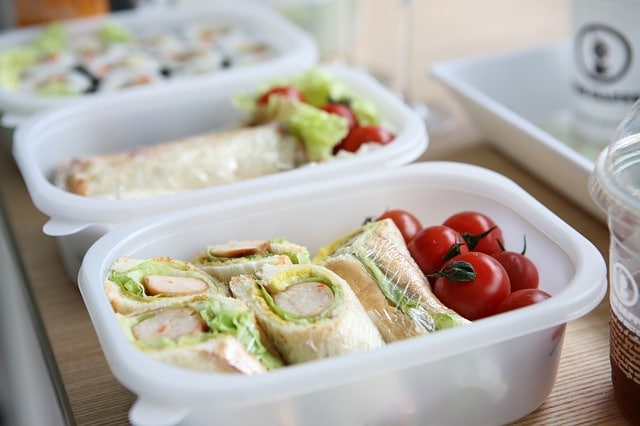 A plastic container filled with sandwich wraps containing lettuce and crab sticks, with cherry tomatoes on the side—ideal budget-friendly foods for departure-day energy. Another container with more wraps and salad is visible in the background.