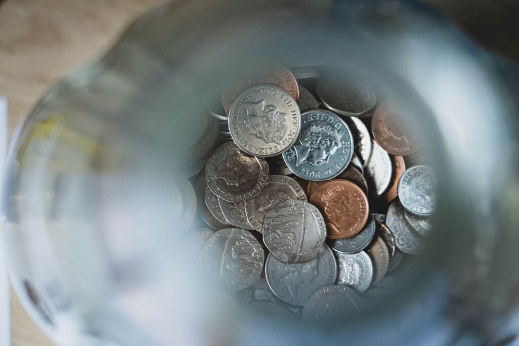A top-down view through the opening of a glass jar filled with assorted coins, including silver and copper ones, illustrates four ways to successfully save more money on a light-coloured surface.