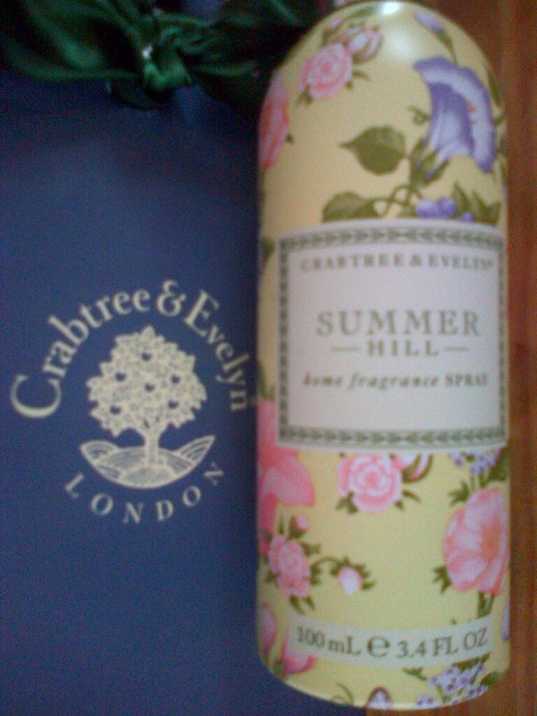 A Crabtree & Evelyn Summer Hill room fragrance spray with a floral patterned bottle—perfect as a gentle sleep aid—is placed on a dark blue surface featuring the Crabtree & Evelyn London logo.