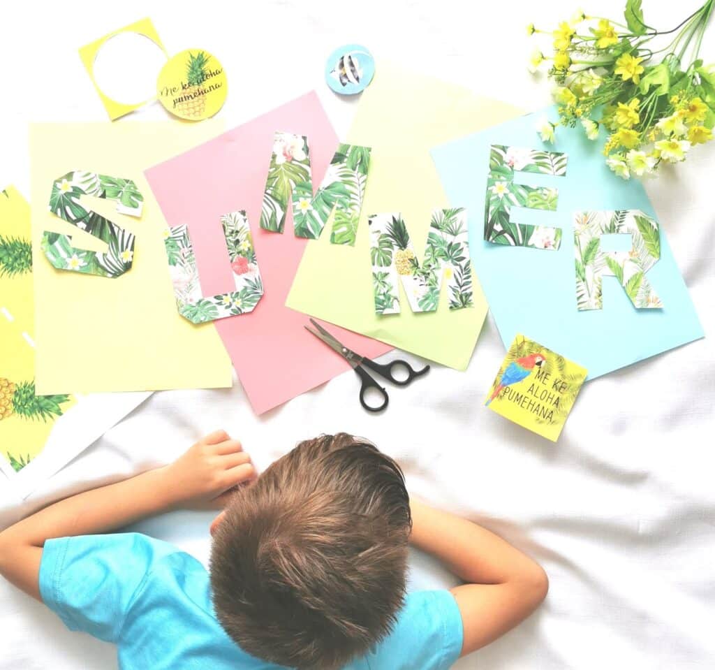 A child lies on a white surface near colourful papers spelling "SUMMER" with floral patterns, surrounded by scissors, yellow flowers, and bright decorations—perfect inspiration for your next Summer Giveaway.