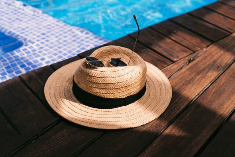 A straw hat with a black band and a pair of sunglasses—classic summer fashion—rest on a wooden decking beside a swimming pool with blue mosaic tiles.